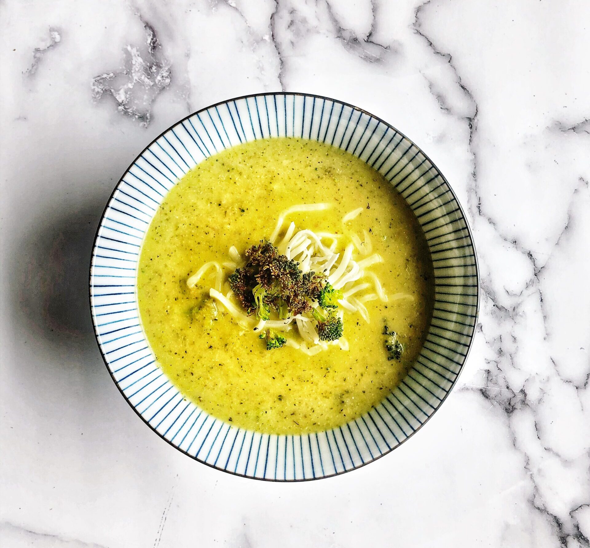 broccoli cheddar soup with crispy broccoli and cheese on top in a blue and white striped bowl on a marble background.