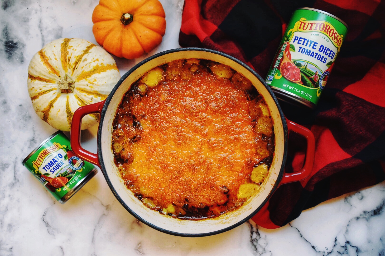 Cheesy Chili Macaroni with a buffalo plaid scarf, marble backgroud, Tuttorosso tomatoes can, and 2 pumpkins.