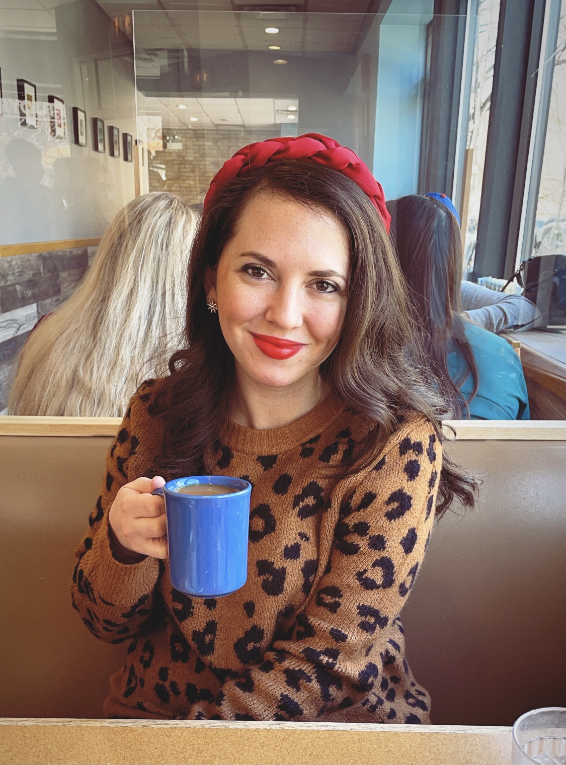 Woman sitting at diner table with blue coffee mug in her hand. She is wearing a cheetah print sweater with long brown hair and a red headband.