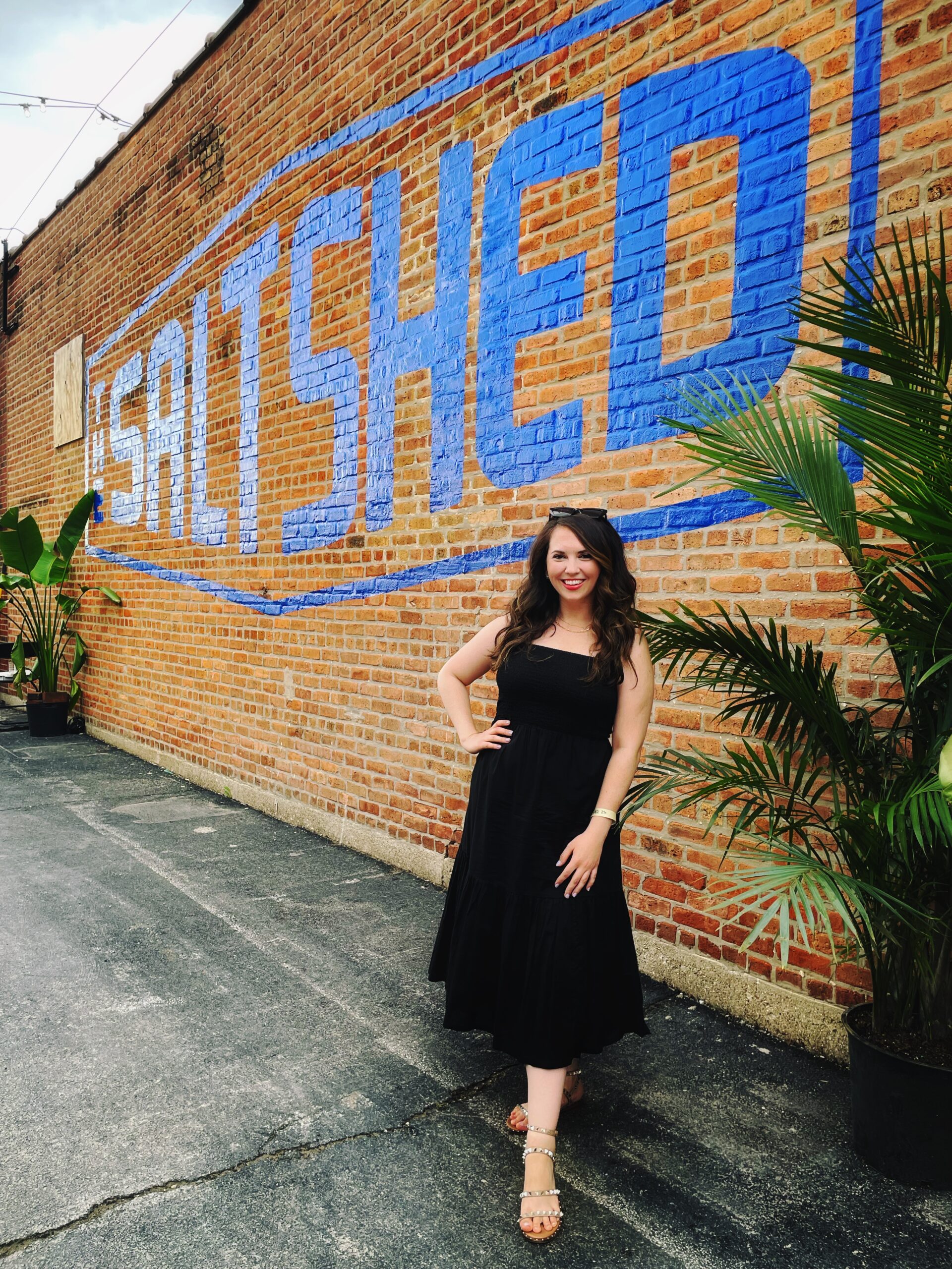 Woman in black dress standing in front of a brick wall that says SALT SHED in blue paint. There is a green plant next to her. She is standing on asphalt ground.