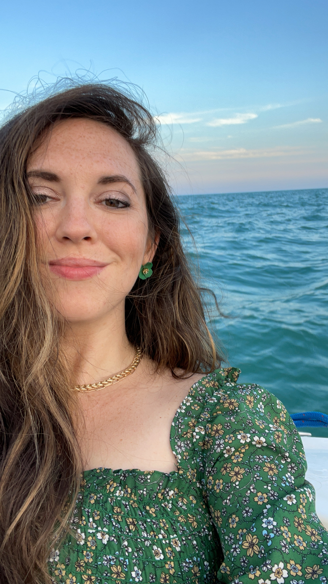 Woman wearing a green shirt with gold jewelry on boat on body of water with sunsetting.