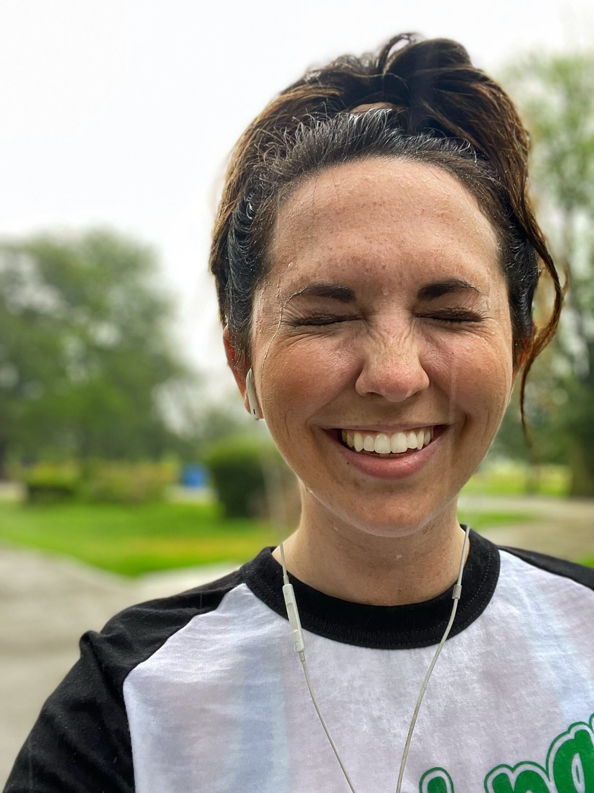 Rain coming down on a woman with brown hair Her eye sare closed and she has brown hair in a ponytail. She is outside with trees in the background. She is wearing a white and black t-shirt with some green print.