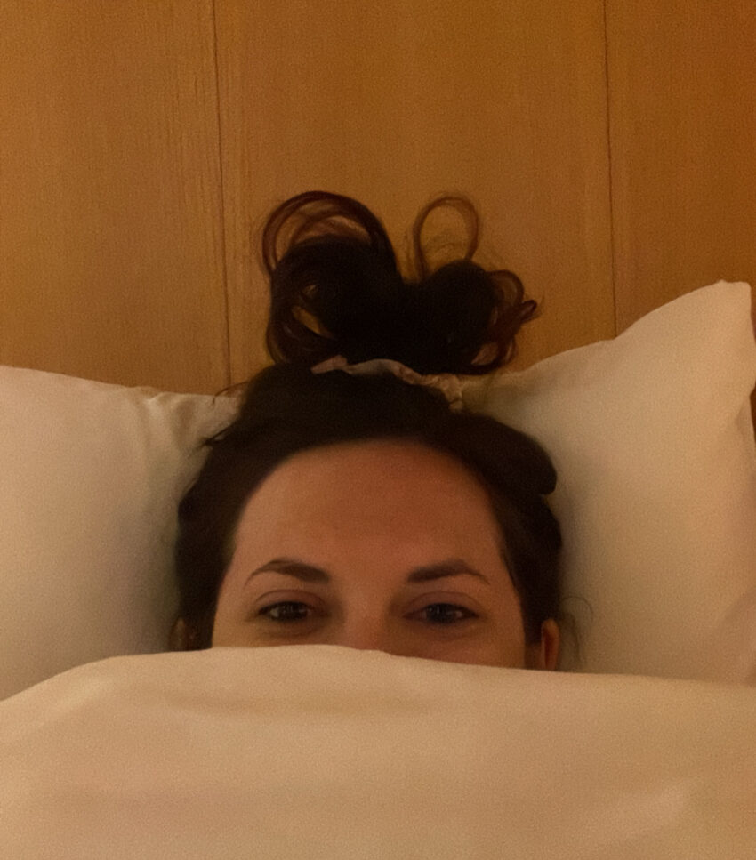 Woman in bed under covers with messy bun. Blanket is white.